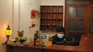 A wooden reception desk with a person seated behind it, a wall-mounted phone, and a shelf with various items. - Hotel Makroz Colonial (Latacunga)
