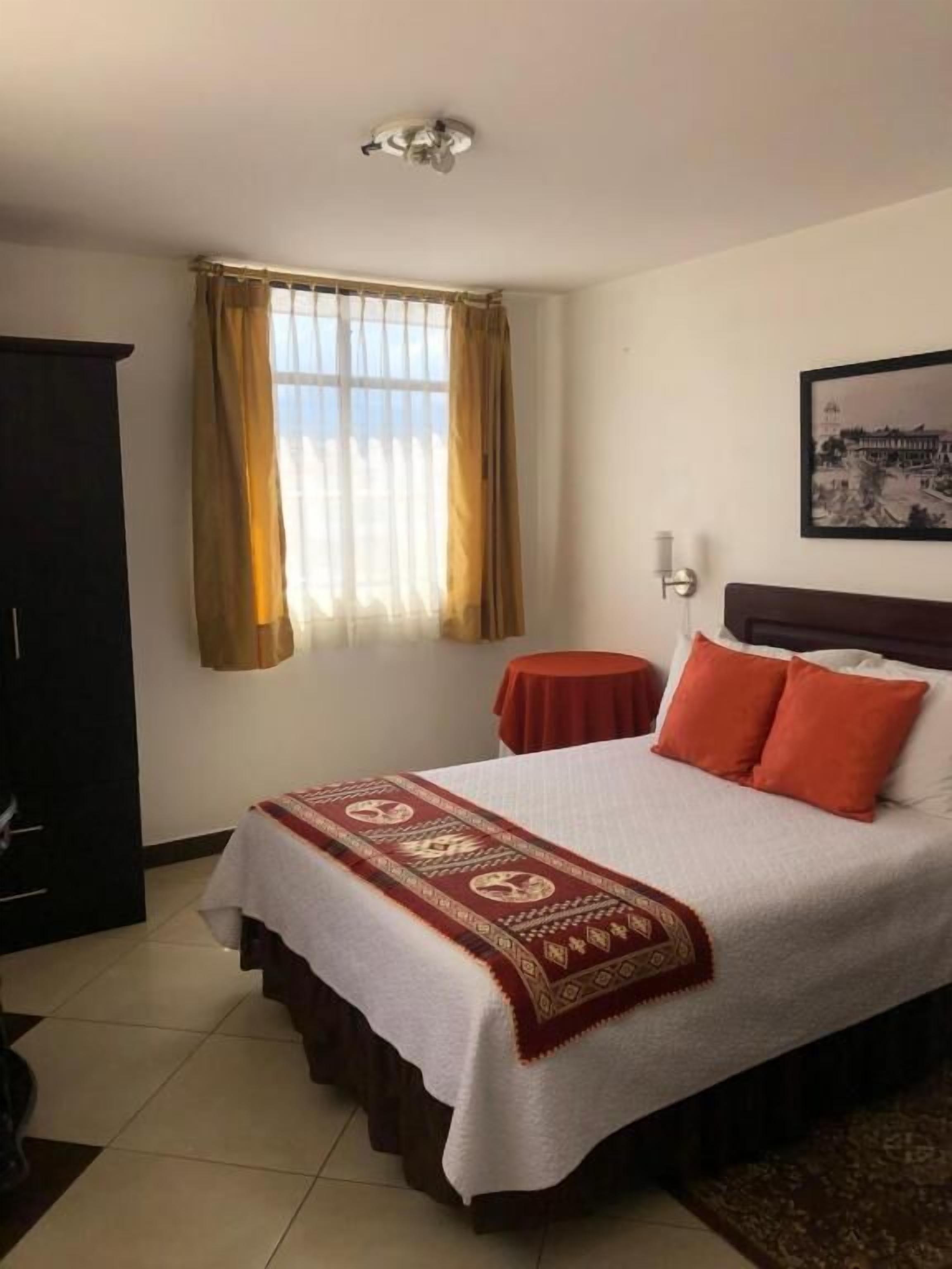 A hotel room with a bed, a small table, a wardrobe, a window with curtains, and a wall-mounted picture.