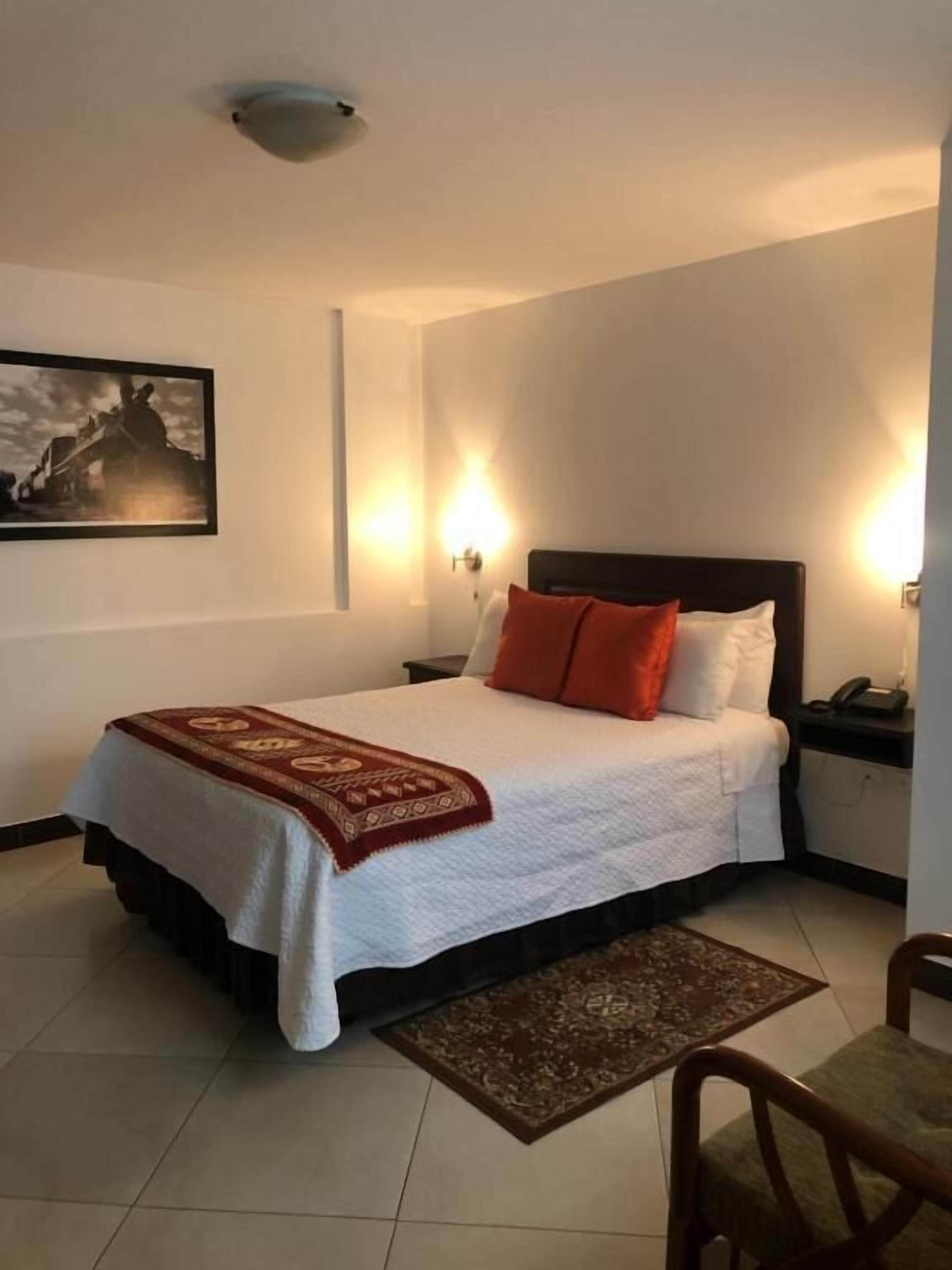 Una cama bien hecha con cubrecama blanco y manta roja, dos lámparas de noche, un cuadro en la pared y una pequeña mesa con una silla.