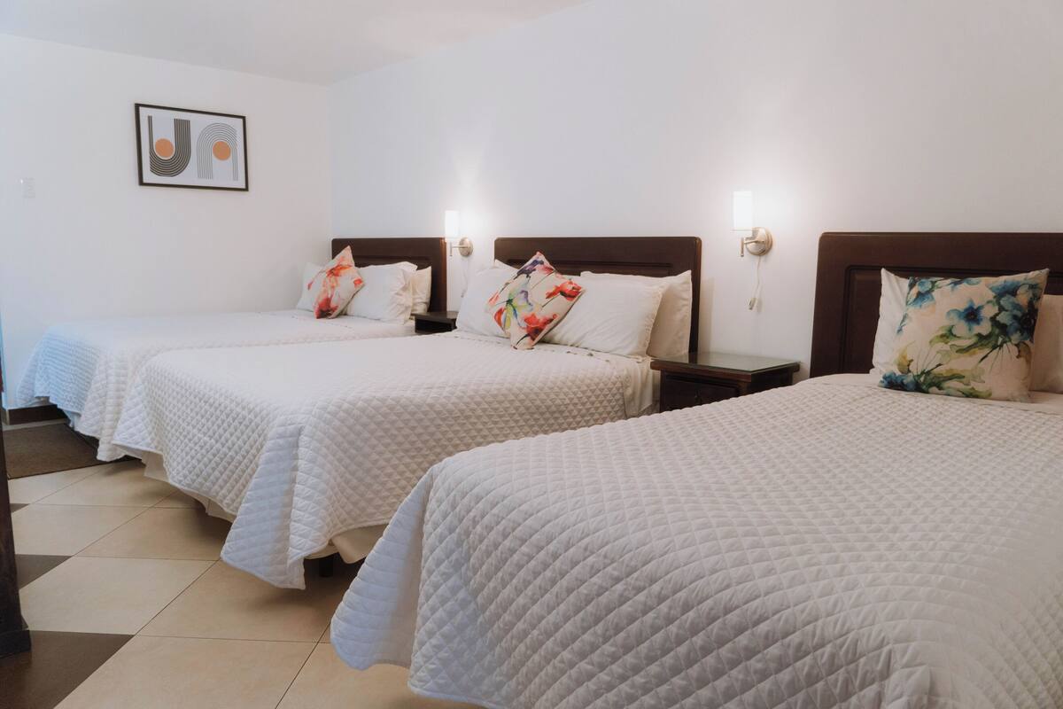 A hotel room with two queen beds, each with a white quilted bedspread and decorative pillows. There is a framed artwork on the wall above the beds and a nightstand with a lamp between them.