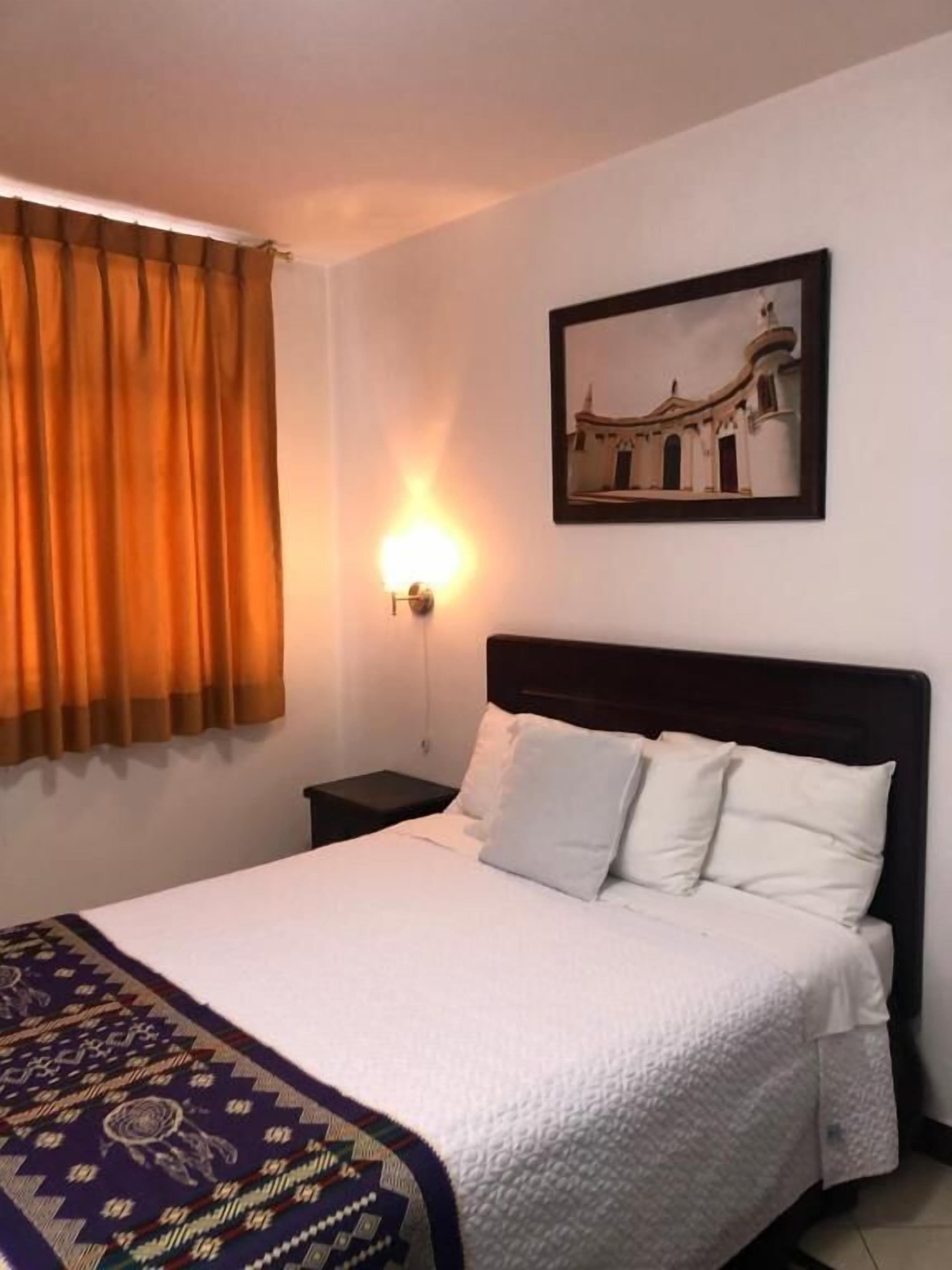 A hotel room with a bed, a nightstand, a wall-mounted lamp, a window with curtains, and a framed picture on the wall.