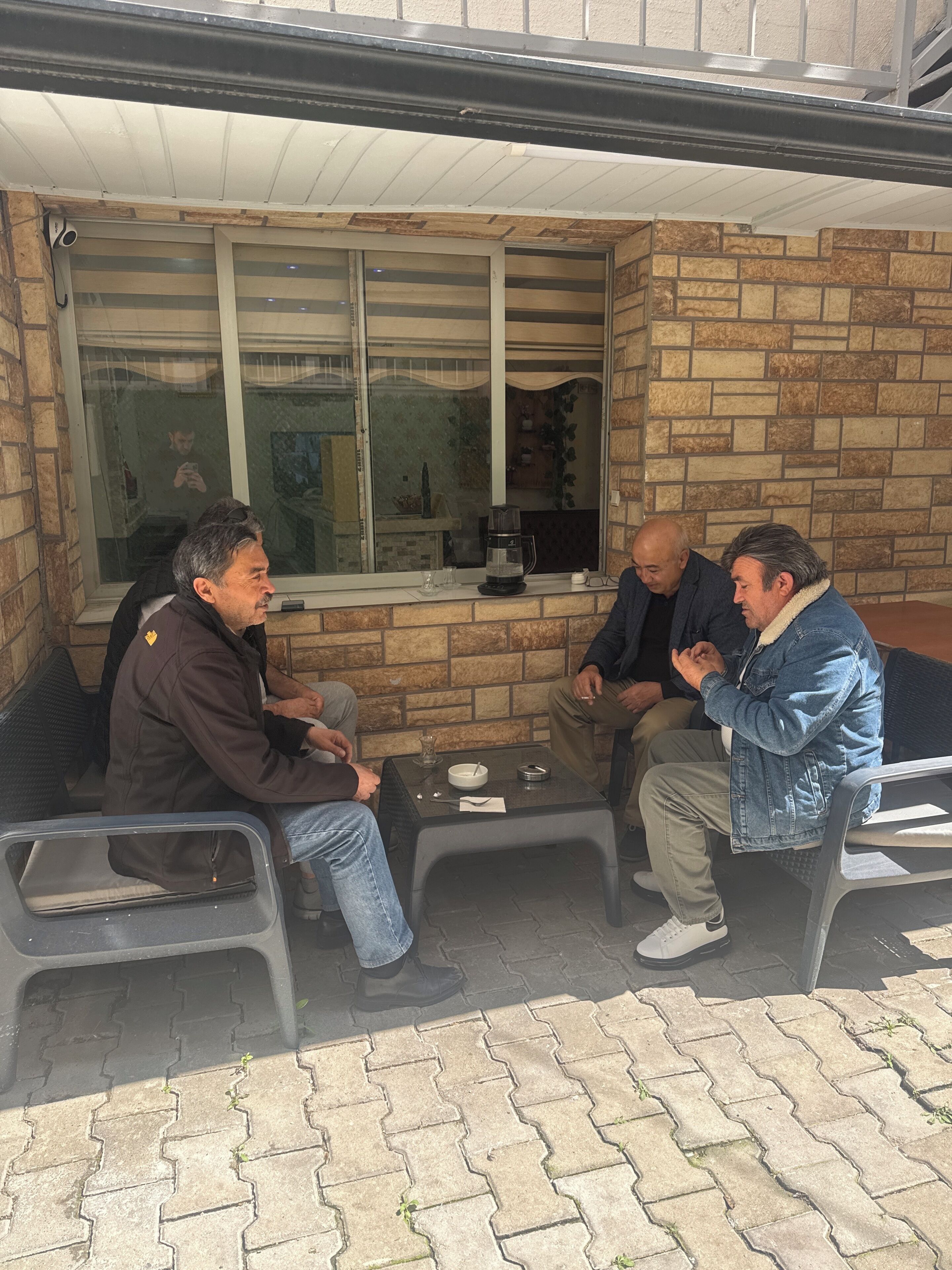 Four men seated at an outdoor table with a brick wall and window in the background.