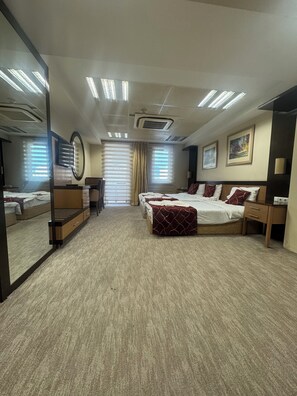 A hotel room with a large bed, bedside tables, a desk, and a mirror. There are windows with blinds, a ceiling air conditioner, and a carpeted floor. - Edremit Körfez Turizm (Edremit)