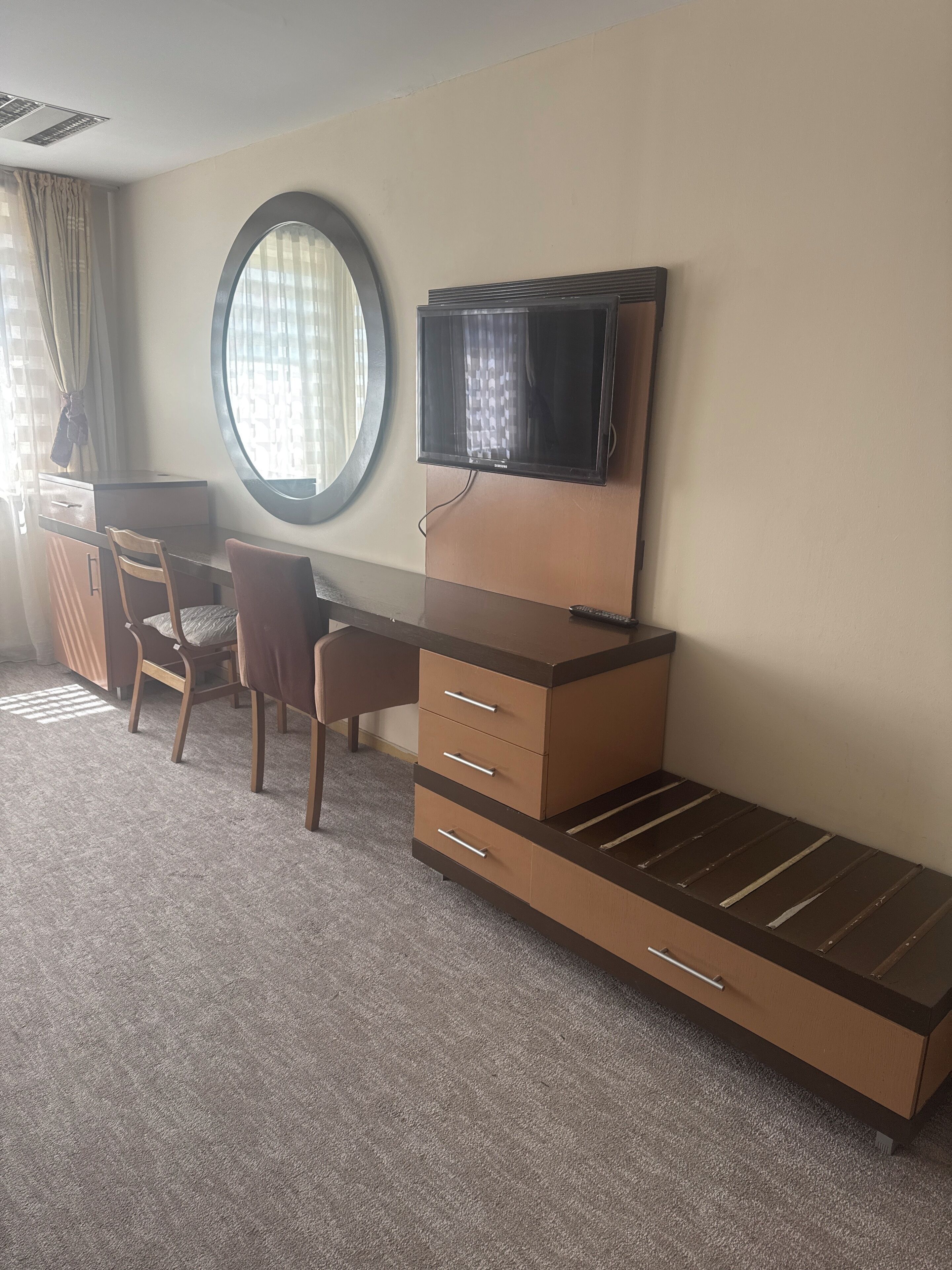 A hotel room with a desk, two chairs, a TV, and a large window with curtains.