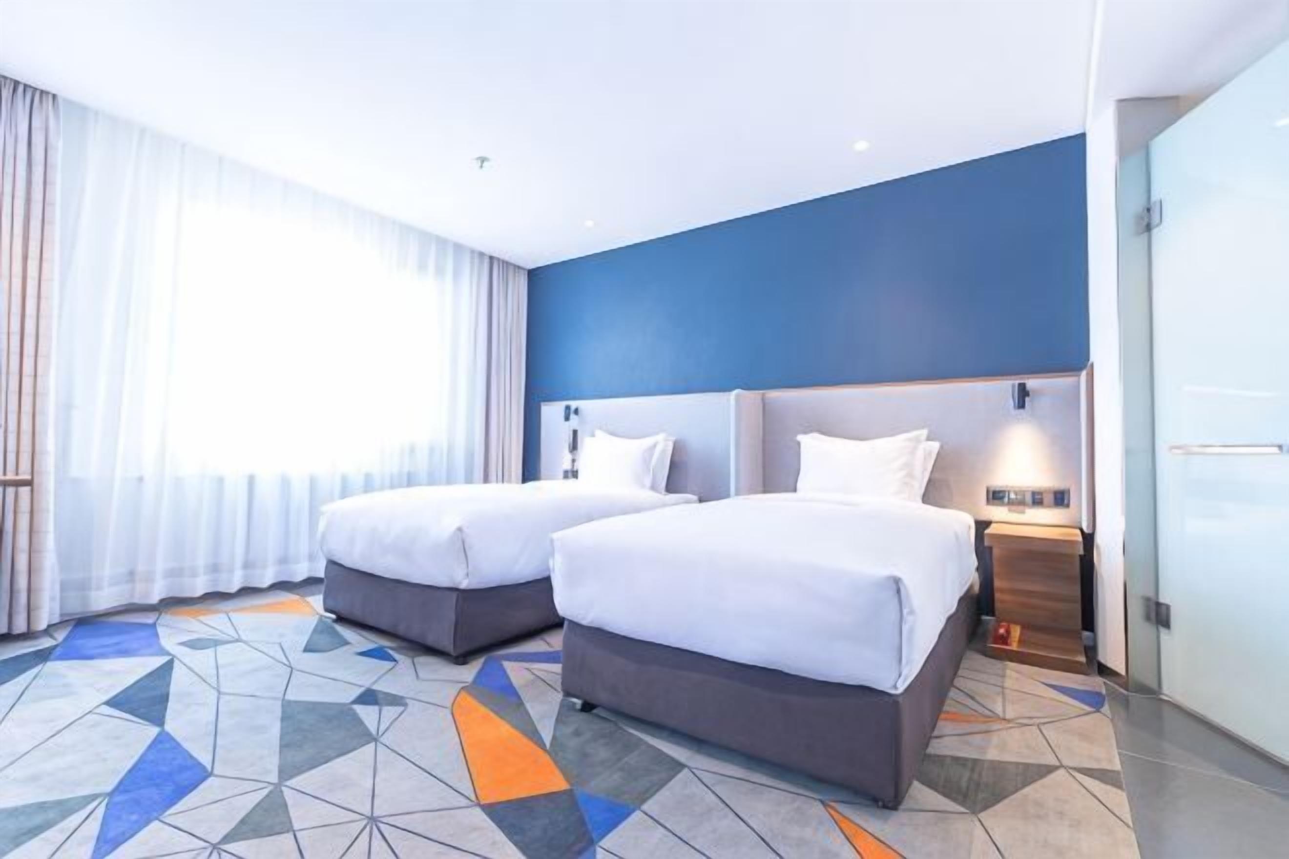 A hotel room with two beds, a large window with curtains, a geometric patterned floor, and a blue accent wall.