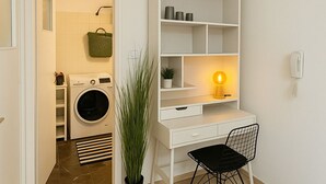 Habitación compacta con un escritorio blanco, una silla, una planta en maceta y una zona de lavandería con lavadora y secadora.