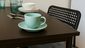 Una mesa de comedor con una taza y platillo azul verdoso, una silla negra perforada e impresiones botánicas enmarcadas en la pared.