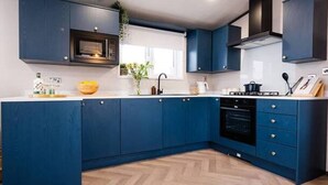 A modern kitchen with dark blue cabinets, a herringbone wood floor, a built-in oven, and a window with white blinds. - Relax at Riverwood (Arbroath)