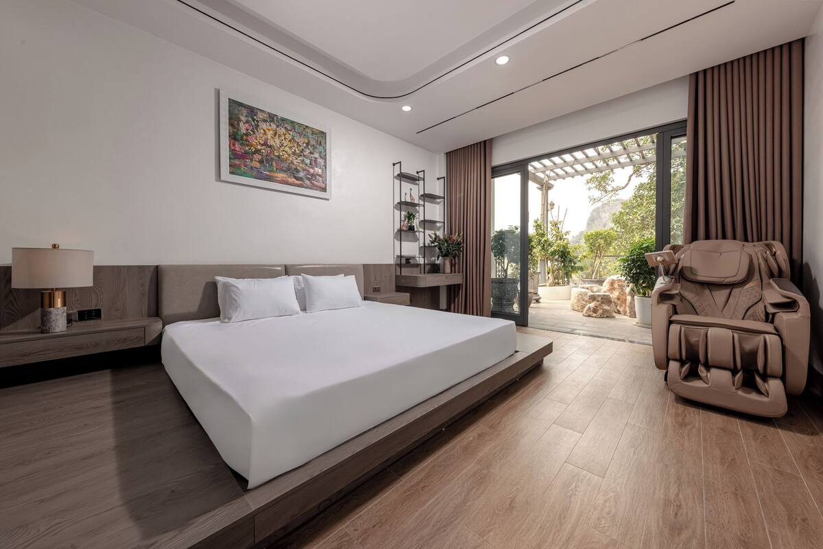 A modern bedroom with a large bed, a massage chair, a small table with a lamp, a bookshelf, and a sliding glass door leading to a balcony.