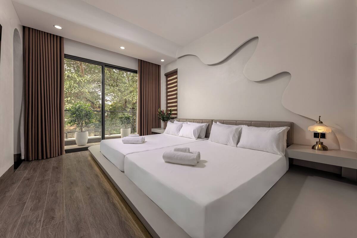 A modern hotel room with a large bed, a bedside table with a lamp, and a view of trees through the window.