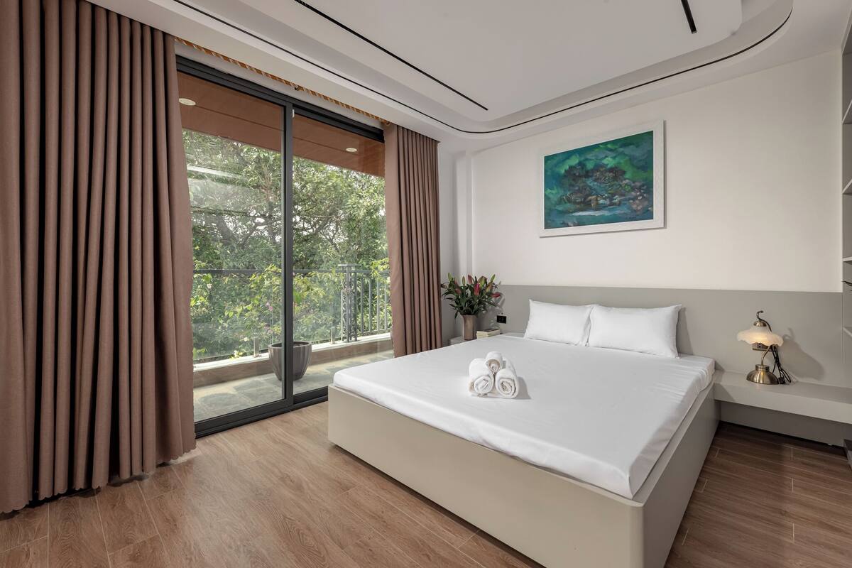 A modern hotel room with a large bed, a view of greenery through a sliding glass door, and a painting on the wall.