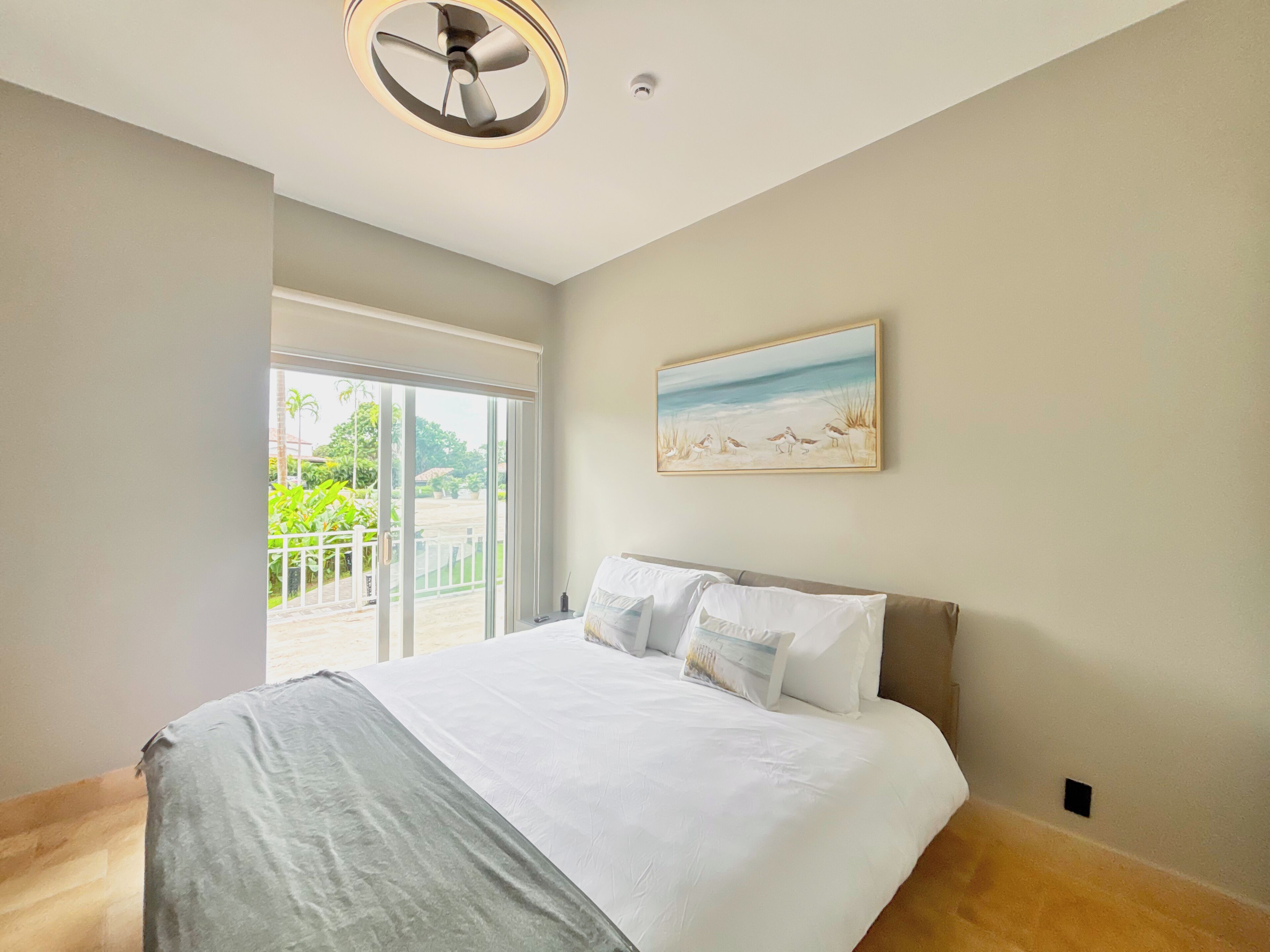 Una camera da letto con un letto grande, una ventola a soffitto, un quadro con degli uccelli al muro e vista su del verde dalla finestra.