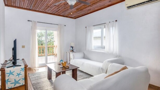 A living room with a wooden ceiling, white sofas, a coffee table, and a TV on a stand. - Tropical Sunset at Sunset Villas in West End, 2 bd Condo  (West End)