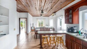 A spacious kitchen with wooden ceiling beams, a central island with stools, and a large window providing natural light. - Tropical Sunset at Sunset Villas in West End, 2 bd Condo  (West End)