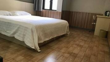 A bedroom with a bed, wooden flooring, a window with curtains, and a small desk with a chair.