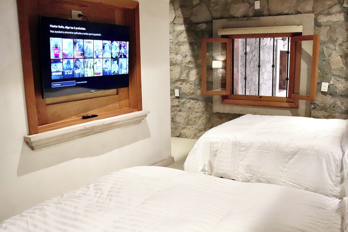 A bedroom with two beds, a television mounted on a wooden shelf, and a stone wall.