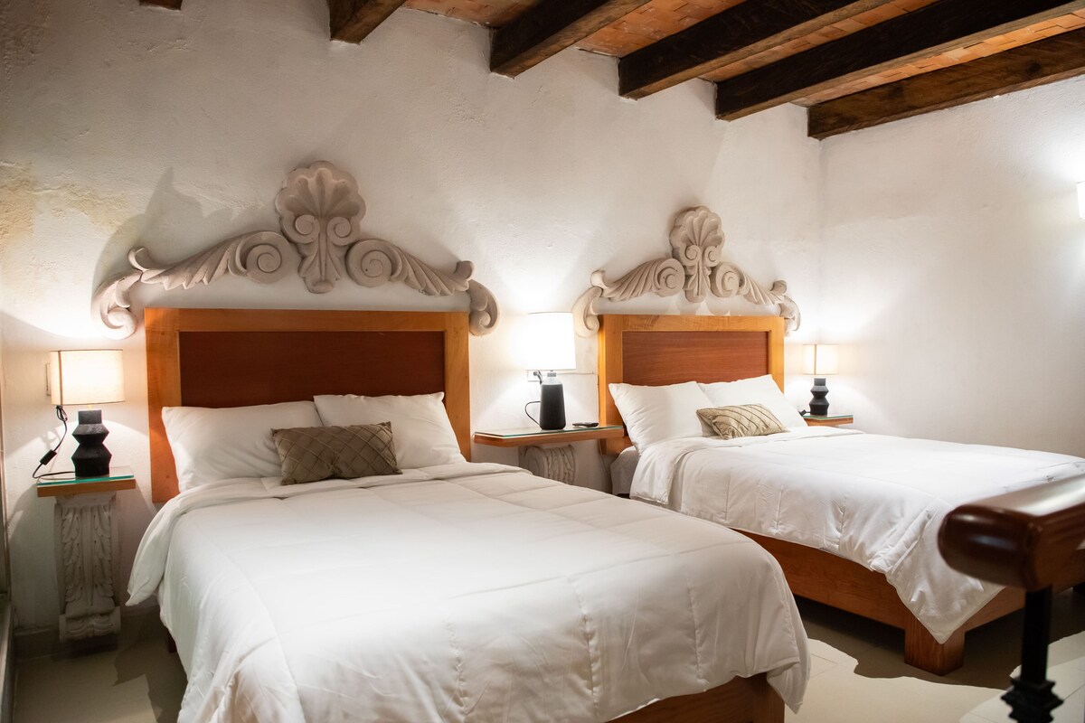 A hotel room with two beds, wooden headboards, white bedding, bedside tables with lamps, and a stone fireplace.
