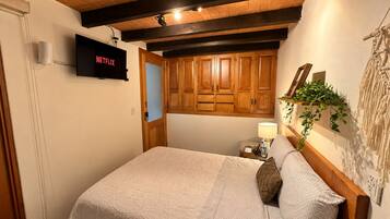 A hotel room with a bed, wooden ceiling beams, a television, a nightstand, a lamp, and a wall-mounted plant.