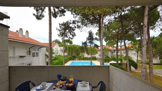 A balcony with a table set for breakfast, overlooking a swimming pool and residential buildings. - Holiday apartment with air conditioning, balcony and pool (Lignano Sabbiadoro)