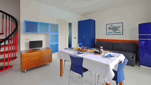 A dining area with a table set for four, a television on a wooden cabinet, blue cabinets, and a red staircase. - Holiday apartment with air conditioning, balcony and pool (Lignano Sabbiadoro)