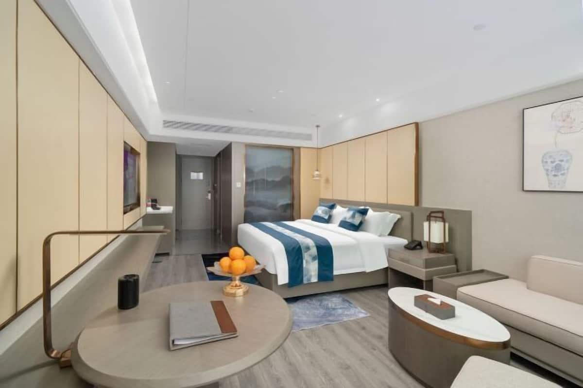 Une chambre d’hôtel moderne avec un lit, une table ronde sur laquelle se trouvent un livre et un panier de fruits, un canapé et une table de chevet avec une lampe.