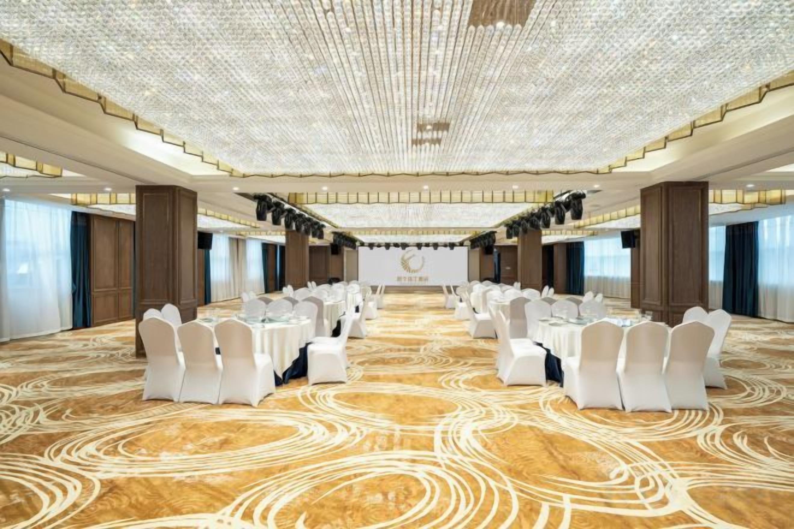Une grande salle de conférence dotée de tables rondes et de chaises blanches, d’un tapis à motifs et d’un plafond haut orné d’un décor.