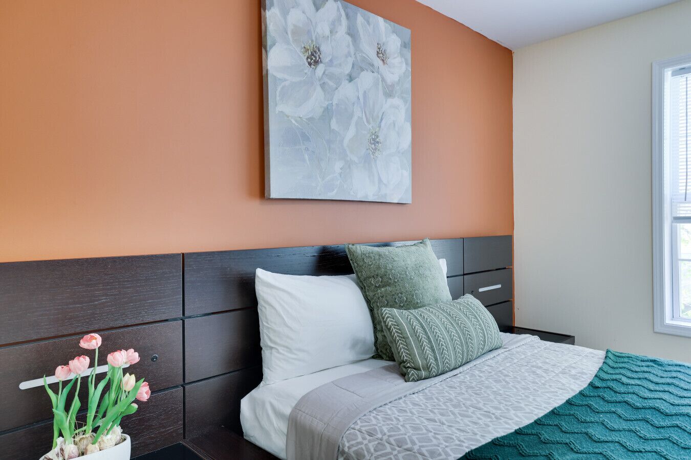 A bedroom with a dark headboard, a bed with white and teal bedding, a green pillow, and a wall-mounted floral artwork.