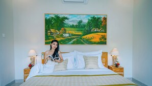 A bed with a patterned bedspread, two bedside tables with lamps, and a painting of a rural landscape on the wall. - Villa Sri Rejeki (Ubud)