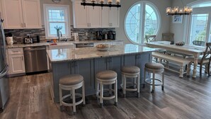 A modern kitchen with a central island, bar stools, and a dining area with a large window. - New Lagoon-Front Beach Home w/ Private Pool, Elevator, & Oceanfront Pool Access (Ocean Isle Beach)