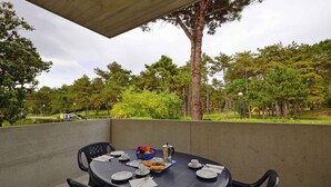 Ein runder Tisch mit Stühlen, Tellern, Tassen und einer Schale mit Früchten und Gebäck auf einem Balkon mit Blick auf einen Park, Bäume und eine Straße im Hintergrund.