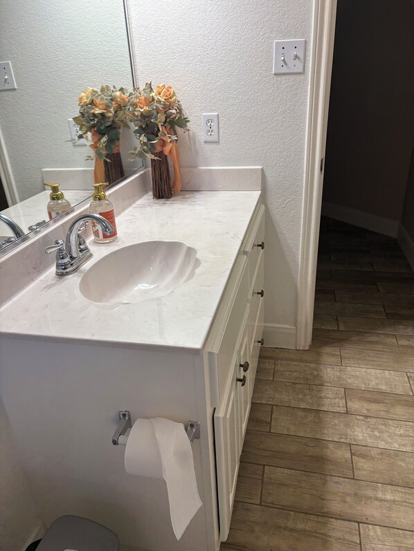 Casa de banho com lavatório branco, espelho, um vaso com flores e suporte para rolo de papel higiénico.