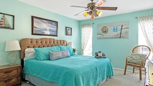 Une chambre à coucher avec une tête de lit capitonnée, un lit avec une literie de couleur turquoise, une table de chevet avec une lampe, une chaise en osier et un ventilateur de plafond.