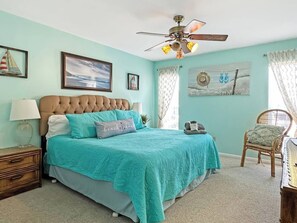 A bedroom with a tufted headboard, a bed with teal bedding, a nightstand with a lamp, a wicker chair, and a ceiling fan. - Oceanfront 2 BR Villa-Pool-Corner Balcony-King Bd (North Myrtle Beach)