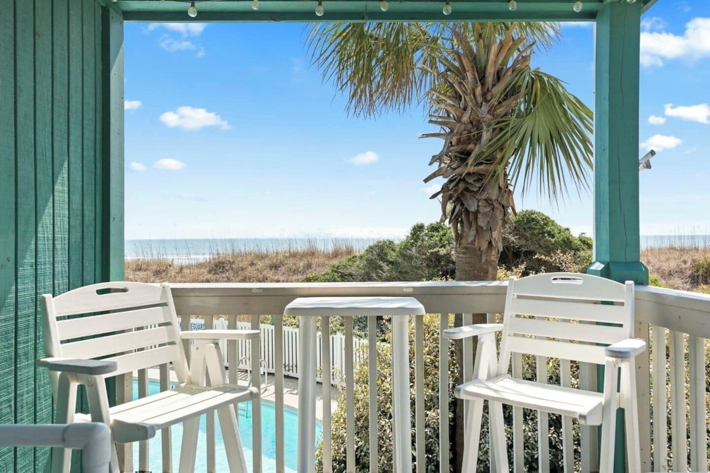 A covered outdoor seating area with white chairs and a table, overlooking a pool and a beach with dunes and a palm tree.