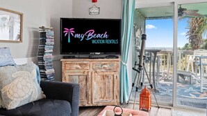 Un salon avec un téléviseur affichant la page « Mes locations de vacances à la plage », une bibliothèque, un canapé et une vue sur l’extérieur.