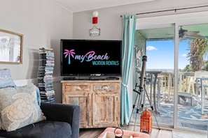 A living room with a television displaying 'my Beach Vacation Rentals,' a bookshelf, a sofa, and a view of the outdoors. - Oceanfront 2 BR Villa-Pool-Corner Balcony-King Bd (North Myrtle Beach)
