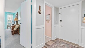 Un couloir avec une porte blanche, une horloge et un tapis menant à une pièce où se trouvent un bureau et une chaise.