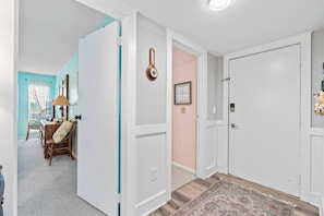 A hallway with a white door, a clock, and a rug leading to a room with a desk and chair. - Oceanfront 2 BR Villa-Pool-Corner Balcony-King Bd (North Myrtle Beach)