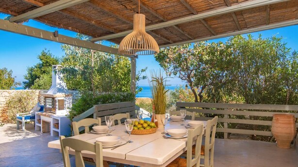 An outdoor dining area with a table set for four, surrounded by white chairs, under a wooden pergola with a hanging light fixture. A stone barbecue grill is visible to the left, and a large potted plant is in the background.