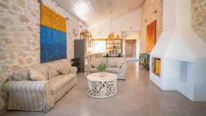A spacious living room with a stone wall, a large painting, a fireplace, and a kitchen area in the background.