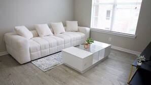A modern living room with a white sofa, a glass coffee table, and a small plant. - Cozy 1BR Magnolia Pool, Gym & Free Parking (Magnolia)