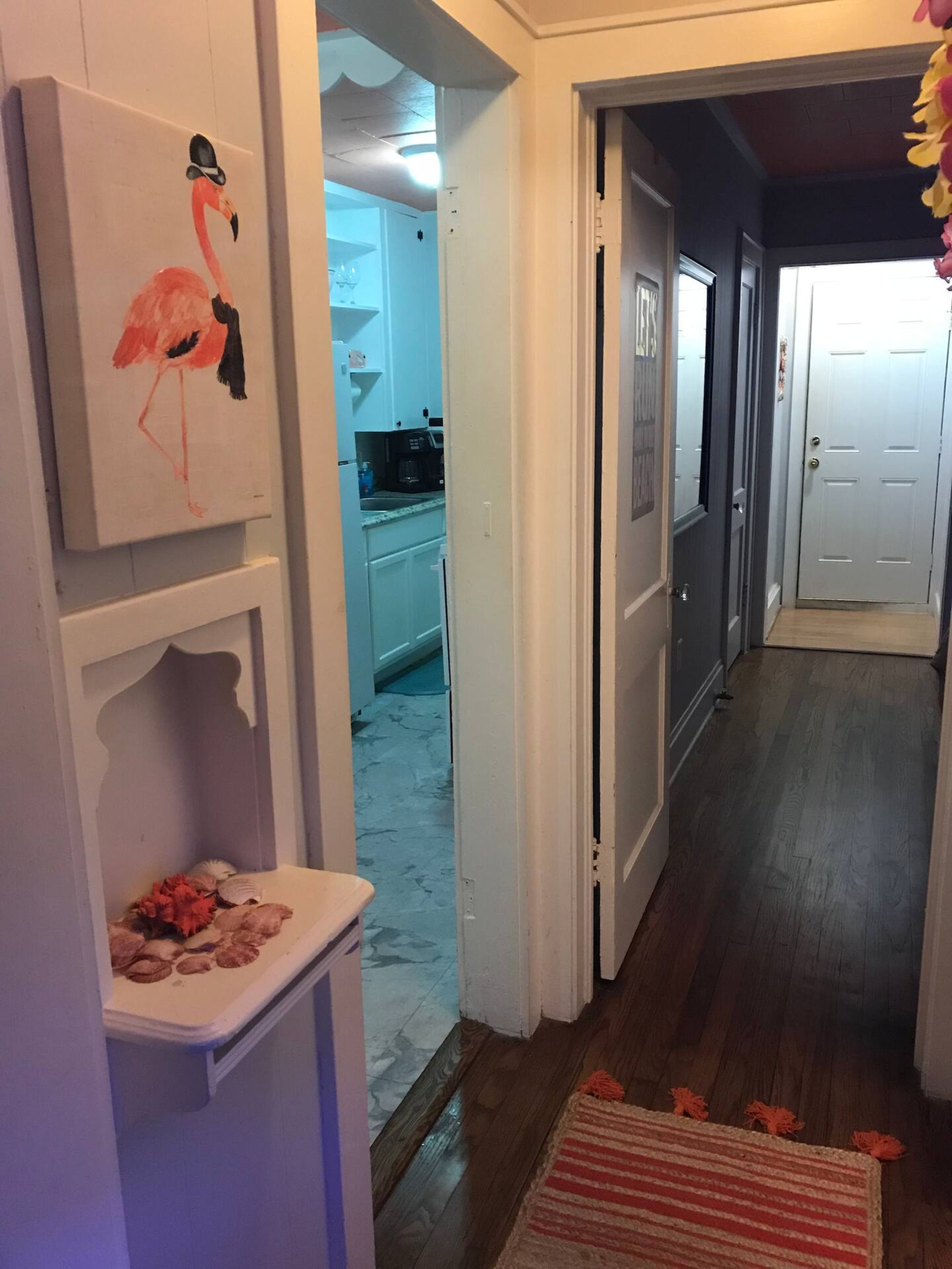A hallway with a kitchen in the background, a door on the right, and a painting of a flamingo on the wall.