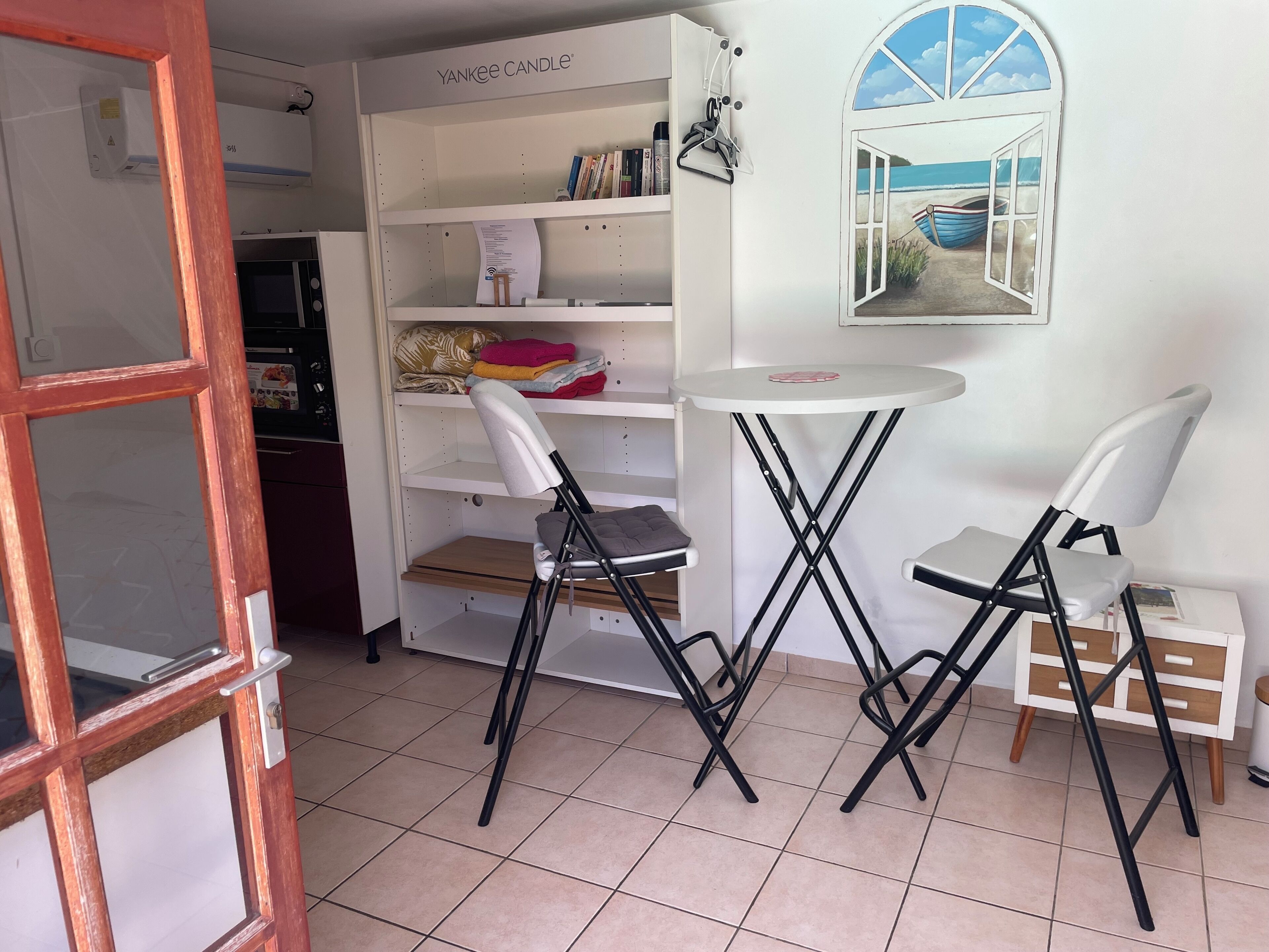 A room with a white bookshelf, a small round table, two black chairs, and a window with a view of the sea.