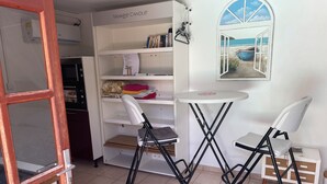 Ein Zimmer mit einer weißen Bücherwand, einem kleinen runden Tisch, zwei schwarzen Klappstühlen, einem Holzkasten und einem Fenster mit Meerblick.