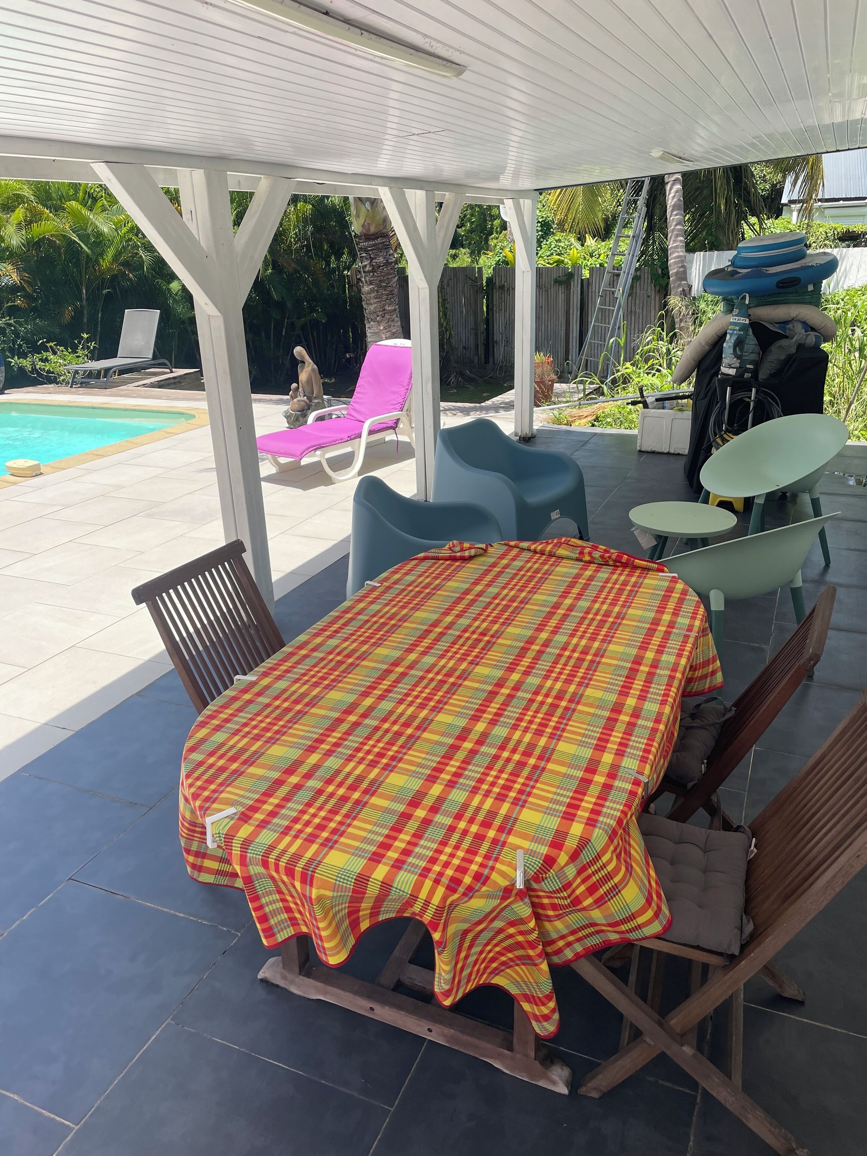 Een overdekt terras met een tafel en stoelen, een zwembad op de achtergrond en een houten omheining.