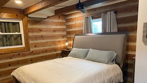A wooden cabin bedroom with a bed, a nightstand, a lamp, and a window with curtains. - Log Cabin at the Farm (Siler City)