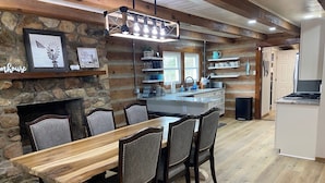 A rustic kitchen with a stone wall, wooden beams, and a dining area with a wooden table and upholstered chairs. - Log Cabin at the Farm (Siler City)