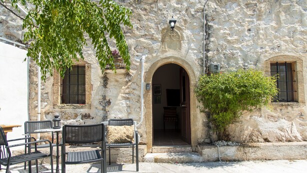Un espace extérieur aménagé avec des chaises et des tables, un bâtiment en pierre doté d’une porte en arc de cercle et un petit arbre.