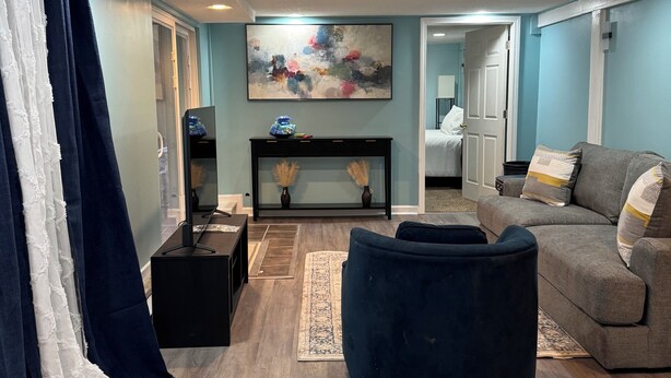 Een woonkamer met een donkerblauwe bank, een rond hocker, een salontafel en een televisie op een dressoir. Aan de muur hangt een groot abstract schilderij en er is een raam met gordijnen.