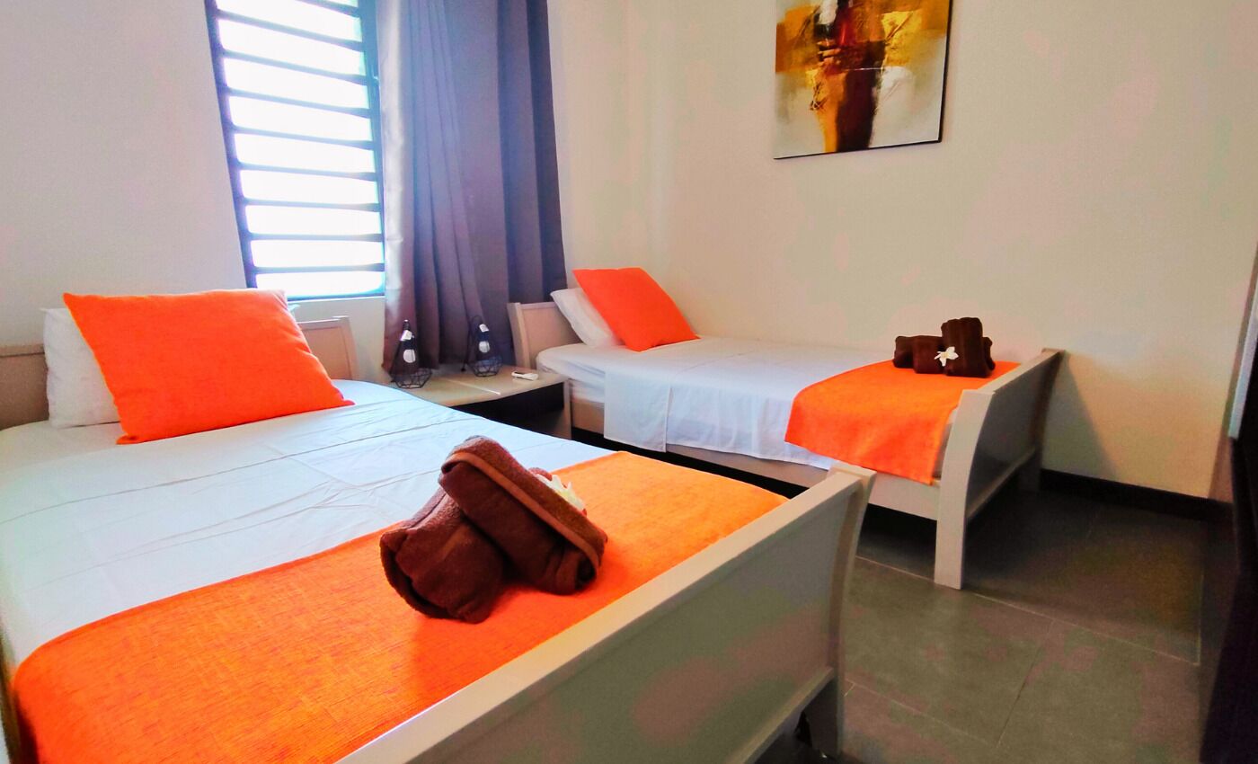 Habitación de hotel con dos camas, cada una con una manta y almohada naranja, un bolso marrón en la cama de la izquierda y una mesita con una botella y vasos.